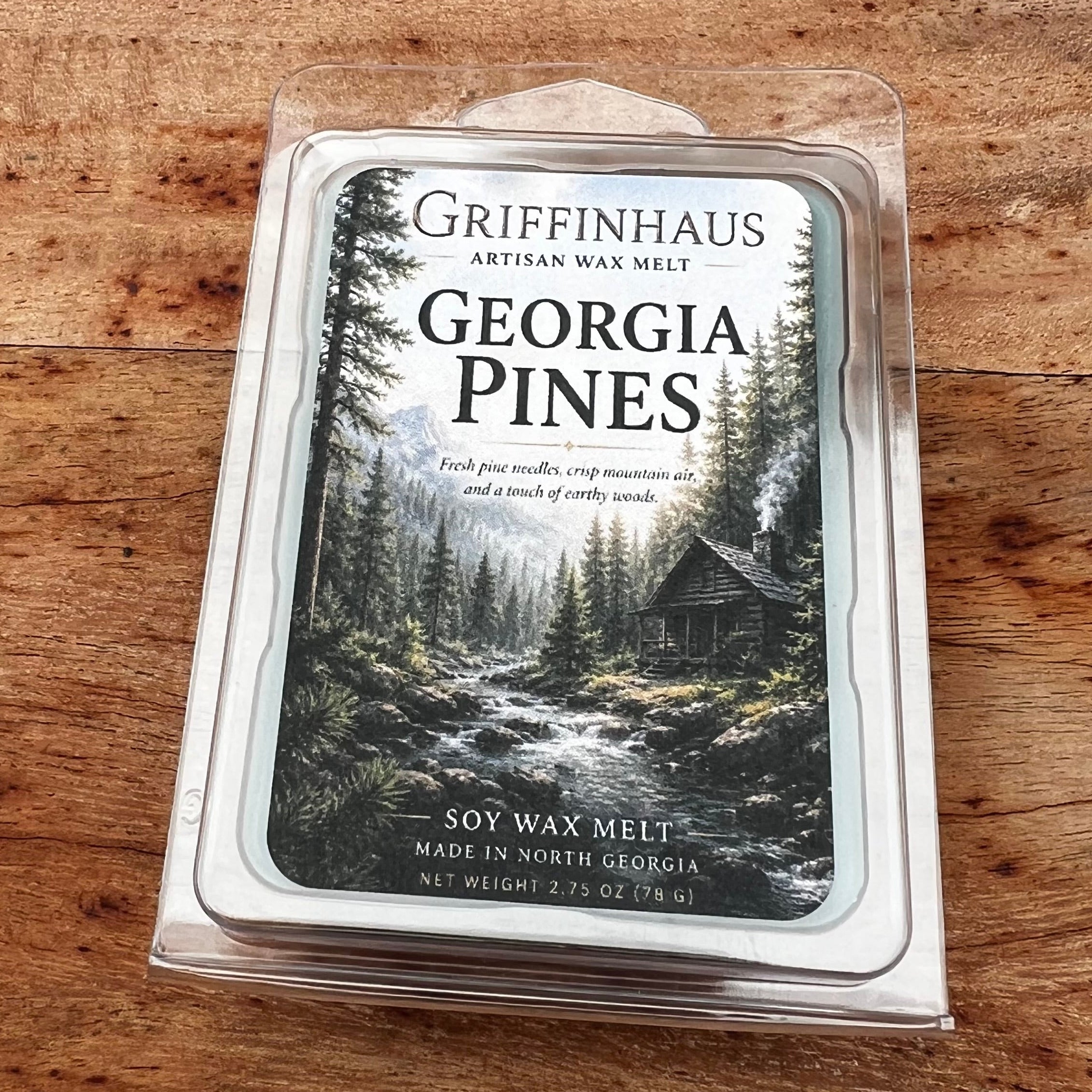 Griffinhaus Georgia Pines artisan wax melt in a clear plastic clamshell package, placed on a rustic wooden surface. The label features a forest cabin scene.
