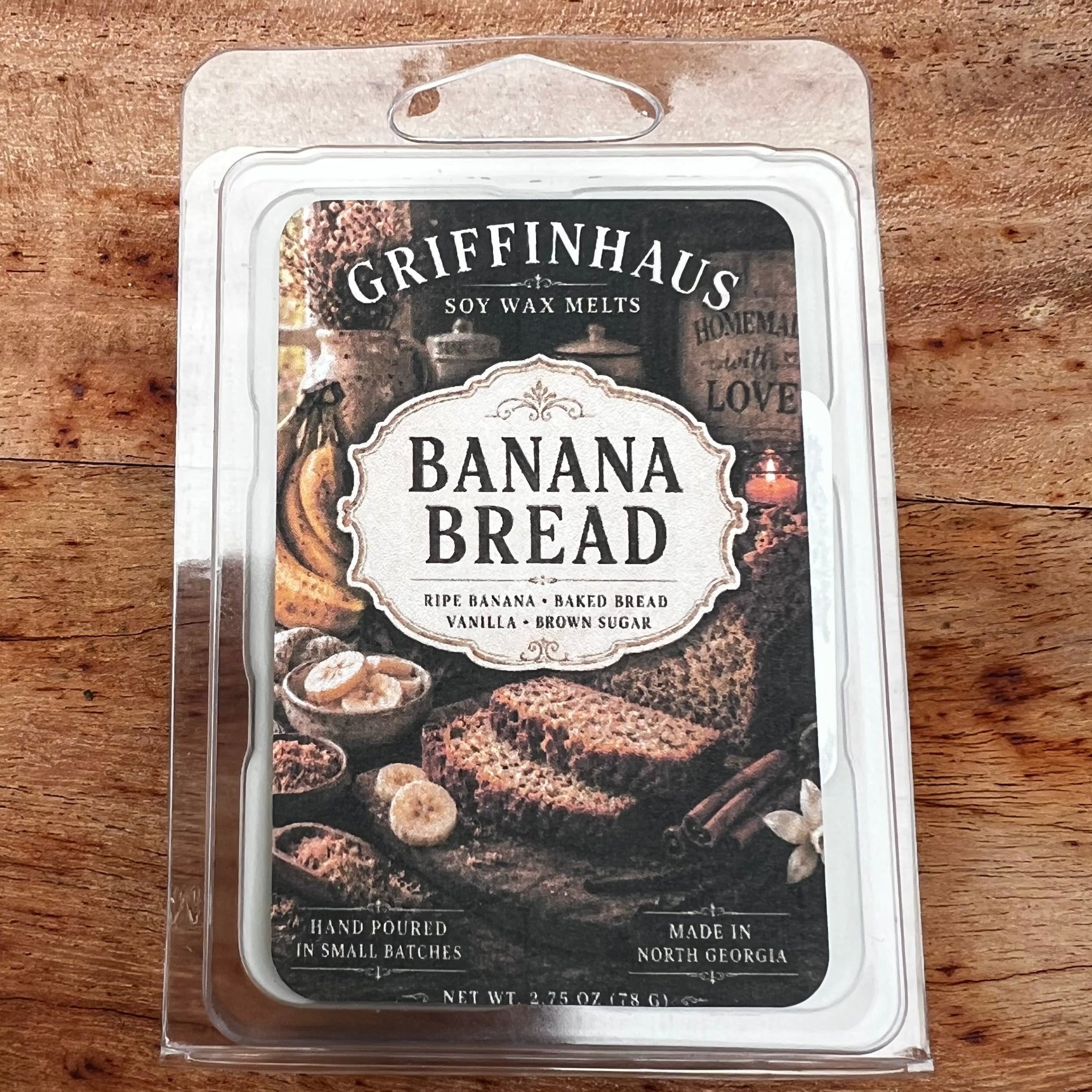 A package of Greenfainhaus Banana Bread scented wax melts in a clear plastic clamshell package, placed on a wooden surface.