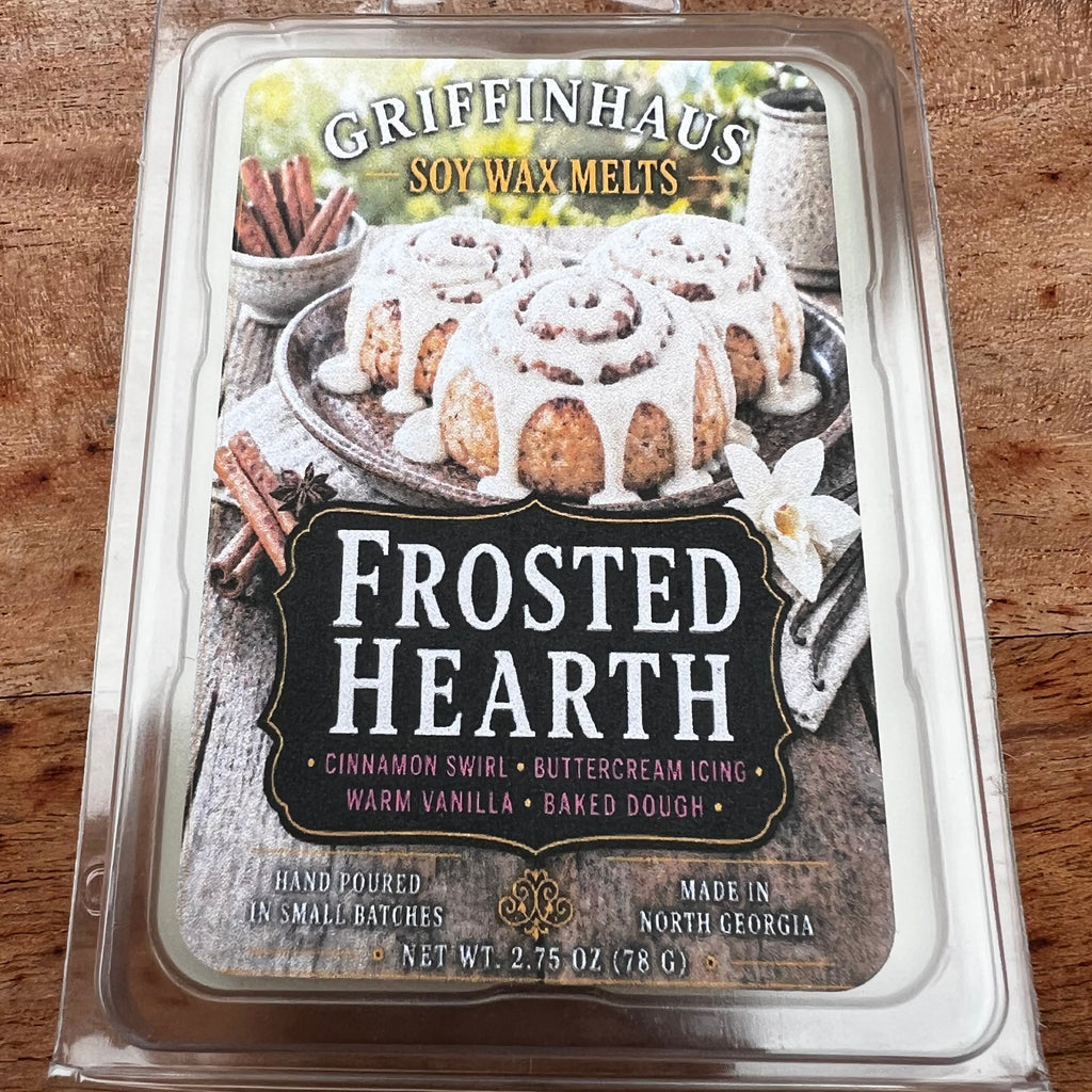 A packaged wax melt product labeled 'Frosted Hearth' in a clear plastic clamshell package, photographed on a wooden surface. The front label shows cinnamon swirls, whipped cream, and the scent name 'Grapefruit + Alms'.