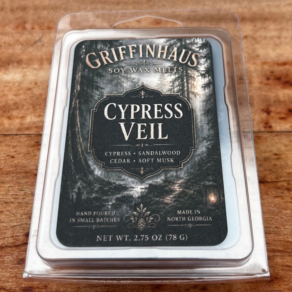 A tin of Griffinghaus Cypress Veil soy wax melts in a clear clamshell package, placed on a wooden surface. The label features a dark, moody forest design with white text.