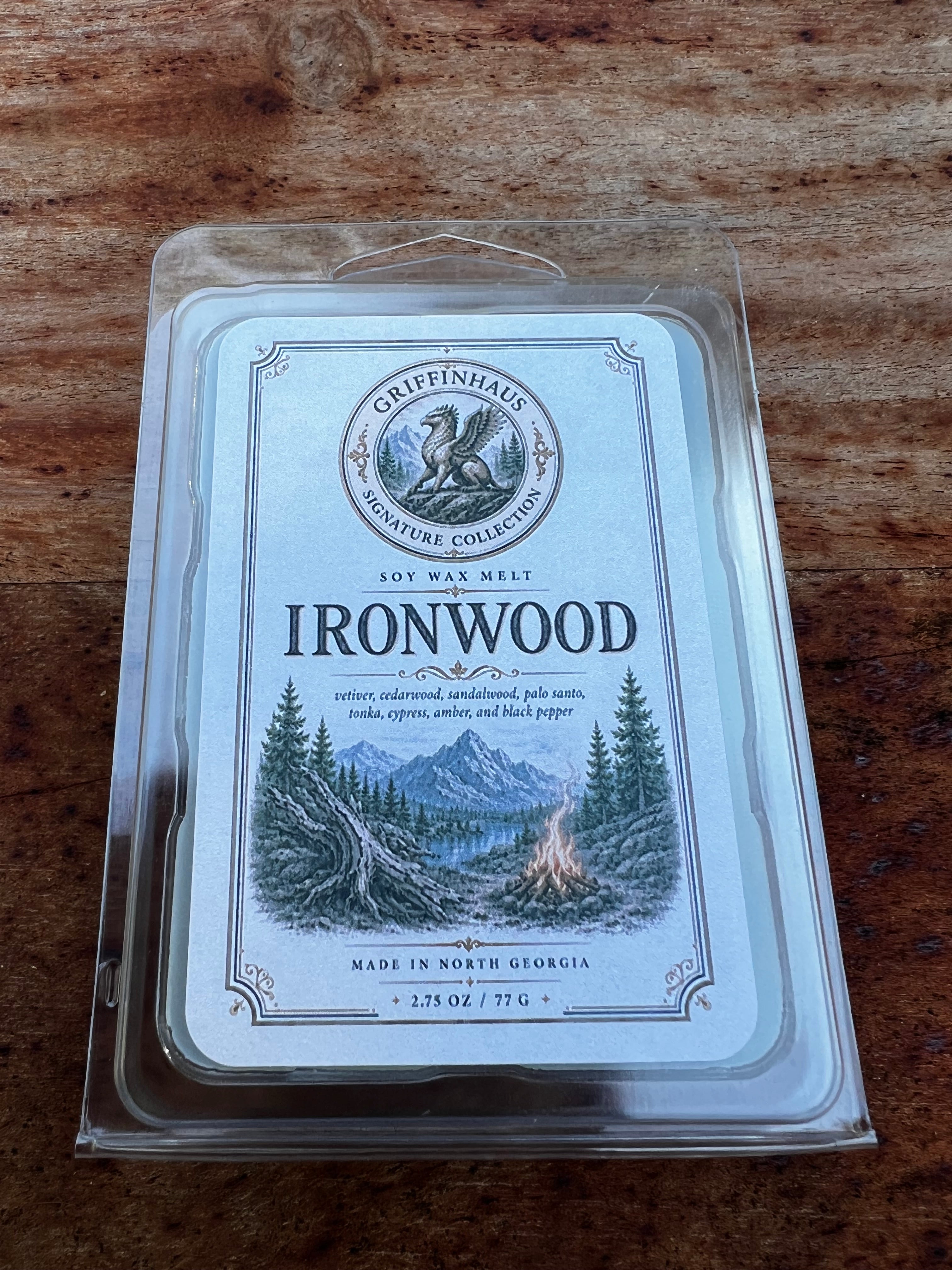 A packaged soy wax melt in a clear plastic clamshell package on a wooden surface. The label reads 'Gripinhal Signature Collection' and 'Ironwood' with an illustrated campfire scene in mountains and trees.