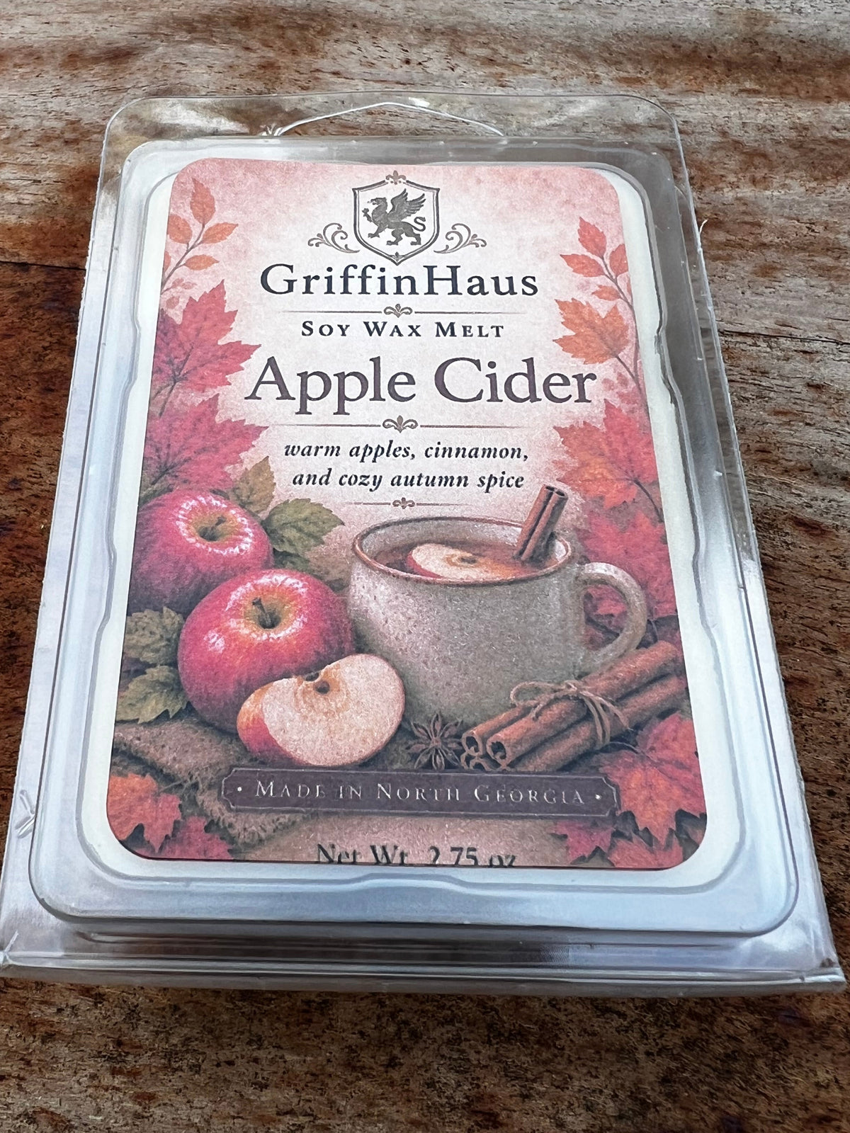 GriffinHaus Apple Cider soy wax melt in a clear plastic clamshell package, resting on a brown wood-grain surface. The label features illustrations of red apples, autumn leaves, and a mug of hot cider.