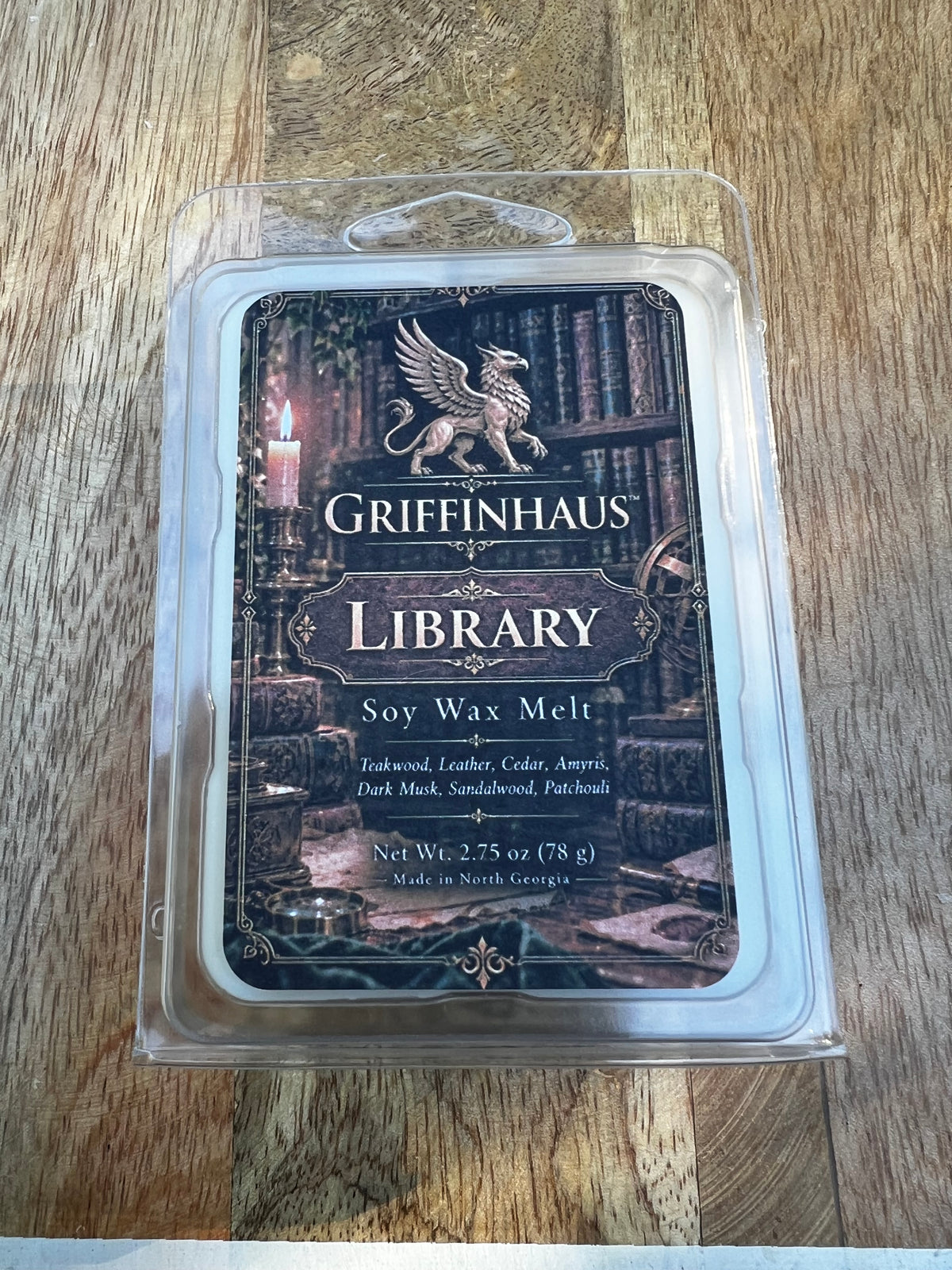 Griffinhaus the Library Soy Wax Melt in a clear plastic clamshell package, placed on a wooden surface. The label features a dark gothic library illustration with a golden griffin and the brand name Griffinhaus.