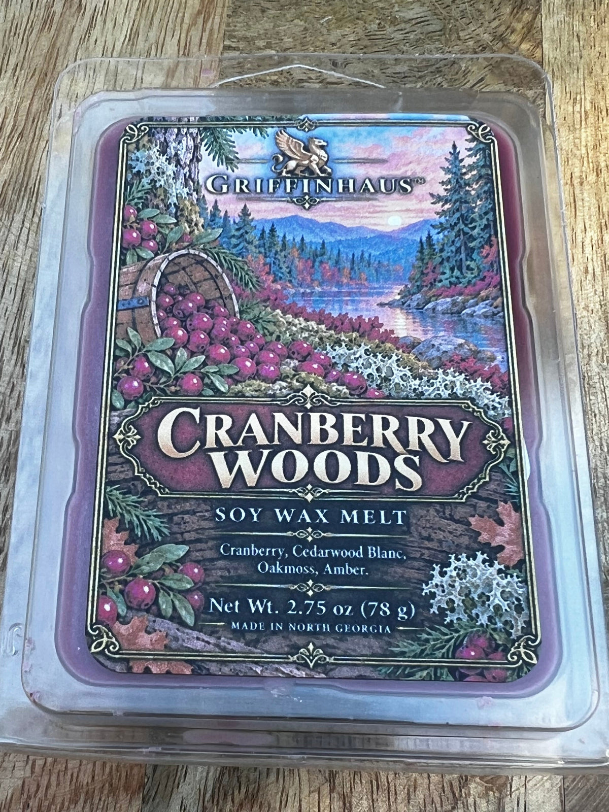 A wax melt package labeled Cranberry Woods by The Artisan Wax Company, placed on a wooden surface. The package is clear plastic with a colorful label showing cranberries, pine trees, and a sunset landscape.