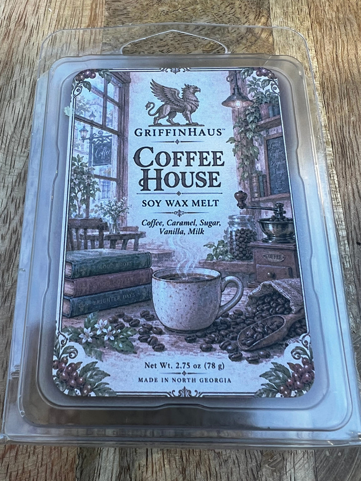 Griffinhaus Coffee House scented soy wax melt in a clear plastic clamshell package, placed on a wooden surface. The label features an illustration of a cozy coffee shop interior with a mug, books, and coffee beans.
