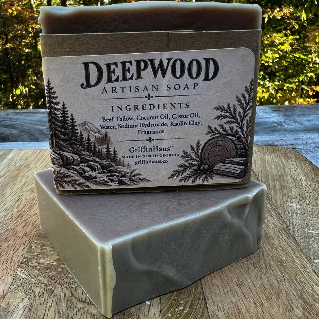 Two bars of Deepwood Artisan Soap stacked on a wooden surface outdoors, with a forest background. The top bar has a kraft paper label reading 'DEEPWOOD ARTISAN SOAP' and 'GriffinHaus'.