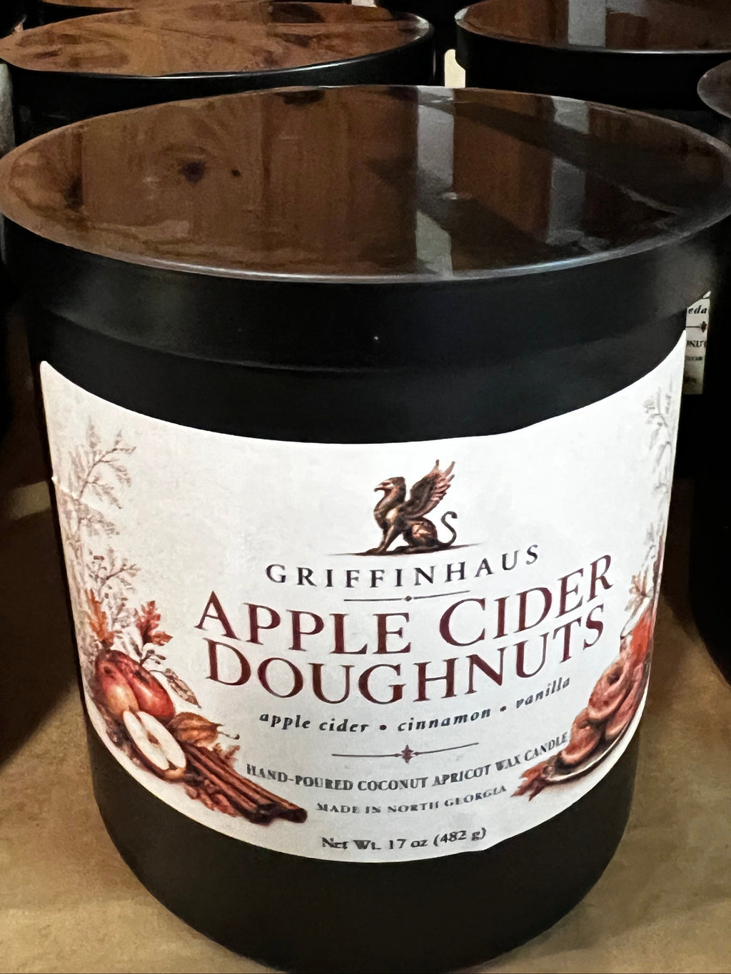 A black jar candle labeled Griffenhaus Apple Cider Doughnuts, placed on a wooden shelf among other candles.