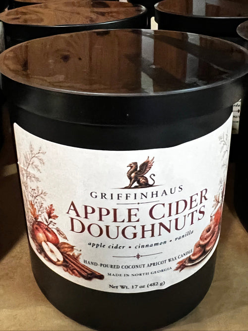 A black jar candle labeled Griffenhaus Apple Cider Doughnuts, placed on a wooden shelf among other candles.